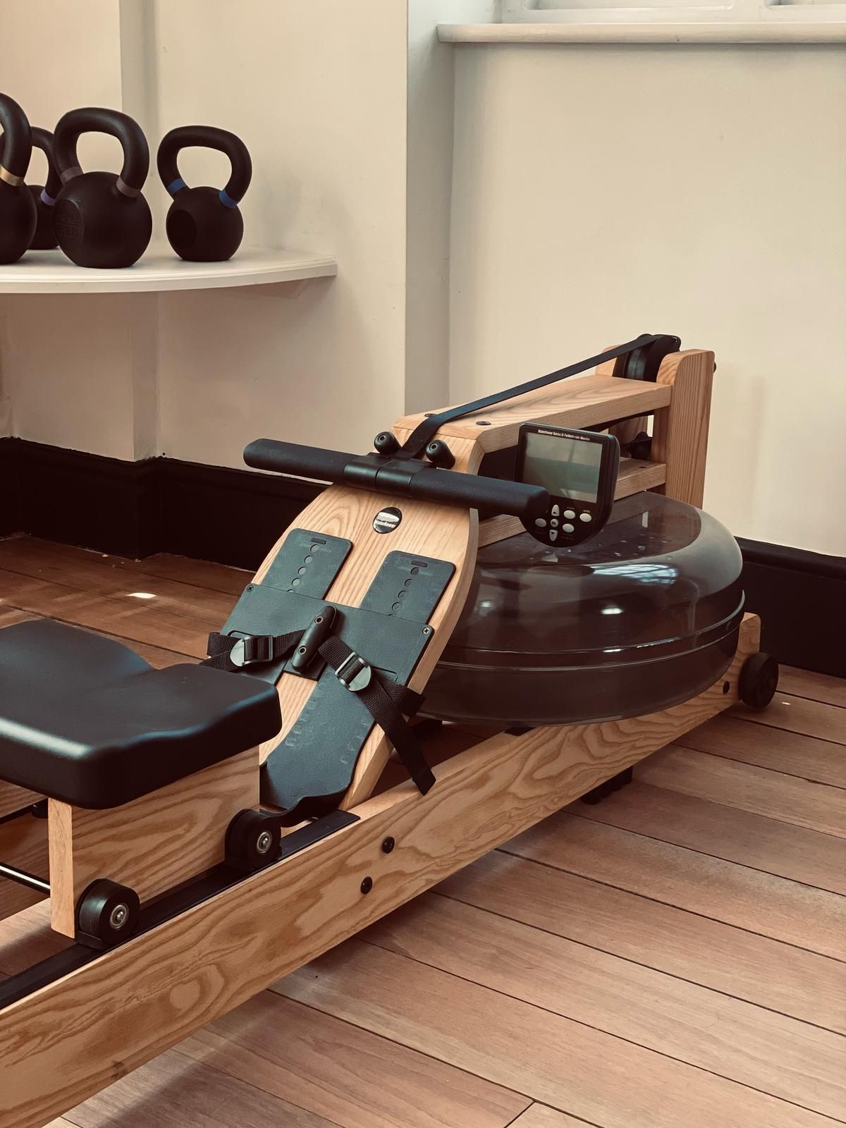 Rowing machine in a gym studio on a wooden floor with kettlebells on a shelf.