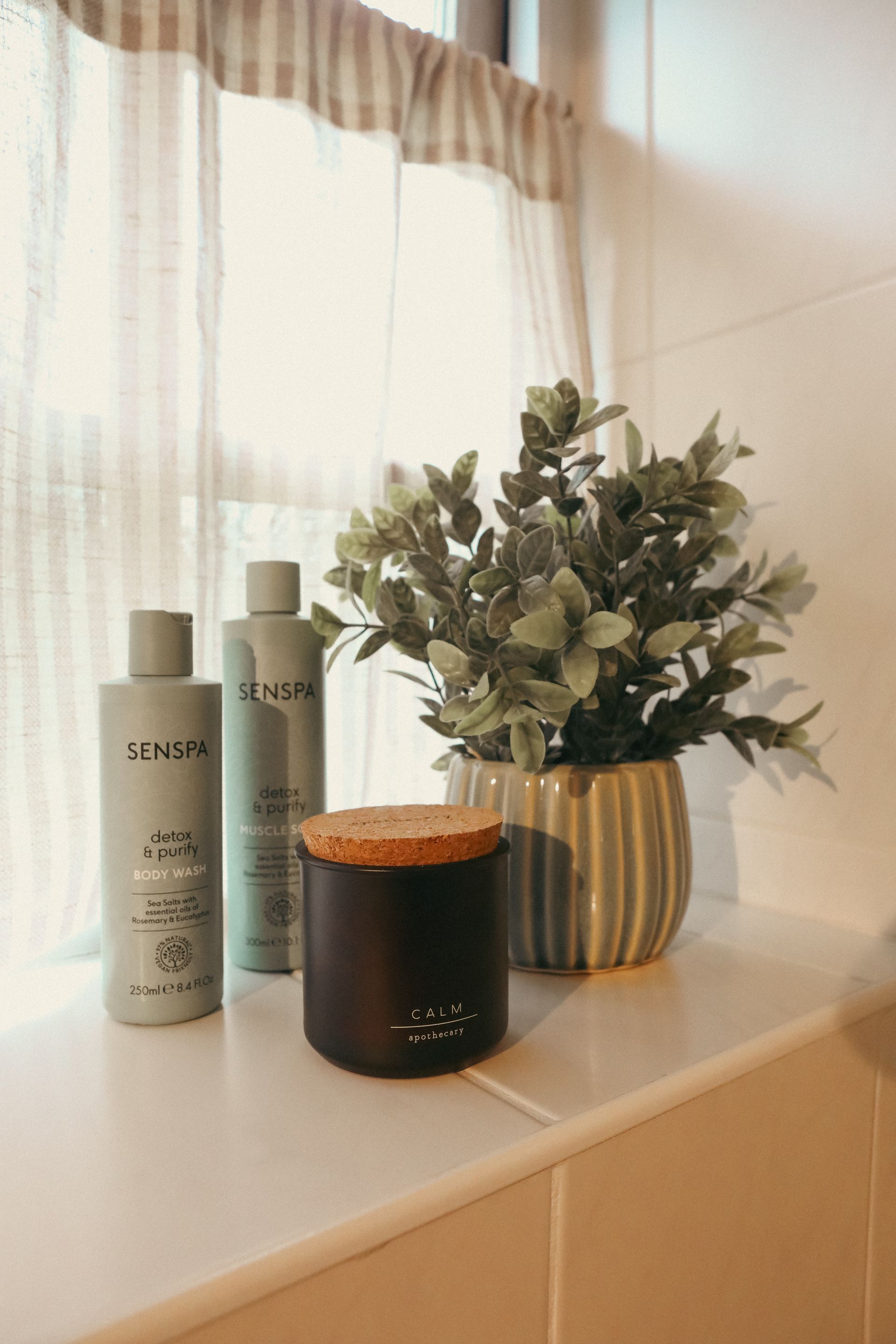 Bathroom vanity with two bottles, a candle, and a striped vase of greenery by a sunlit window