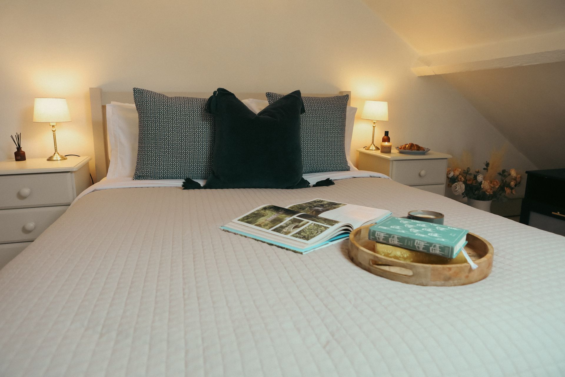 Neatly made bed with white bedding, dark pillows, bedside lamps, and a tray with books and tissues.
