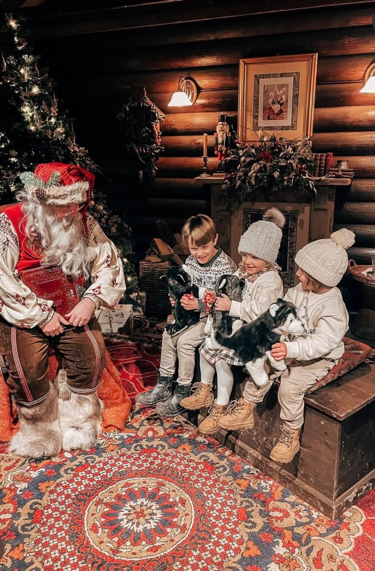 LaplandUK Children in winter clothes sit beside Santa in a cozy log cabin with a decorated Christmas tree.