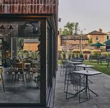 Outdoor café patio with tables and chairs beside a glass-fronted building, overlooking a grassy courtyard