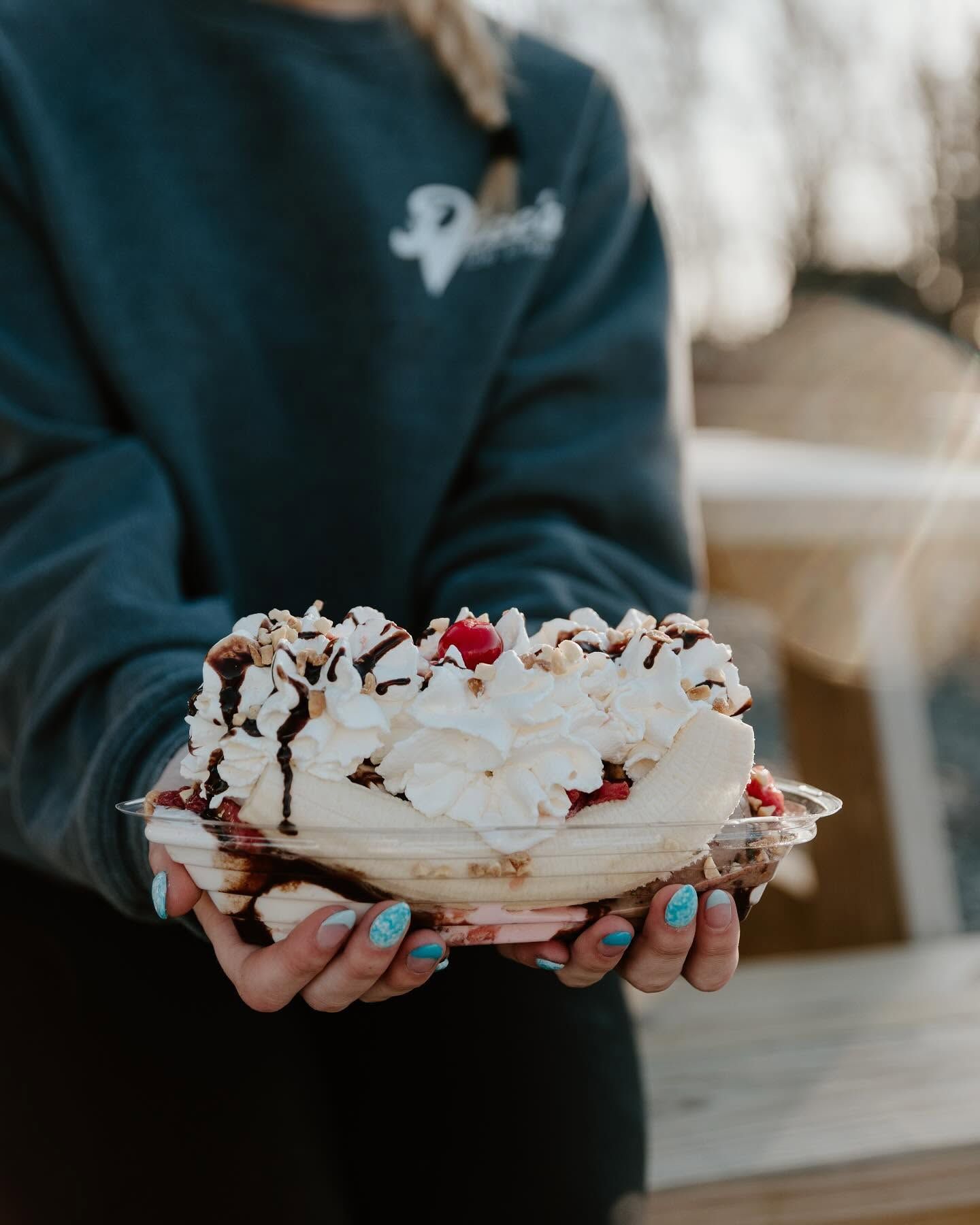 Person holding a layered dessert topped with whipped cream, chocolate drizzle, and cherries outdoors