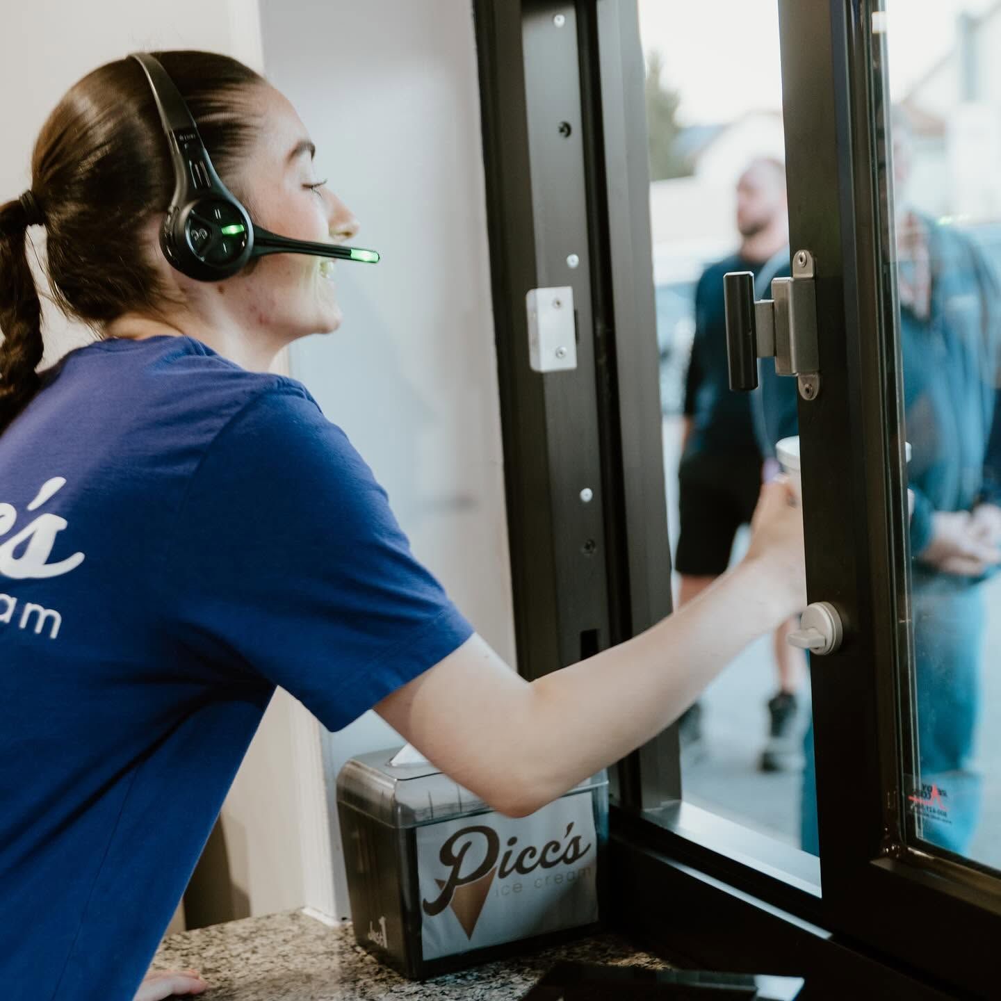 Woman in a Picc's shirt and headset serving a drink through a drive-thru window