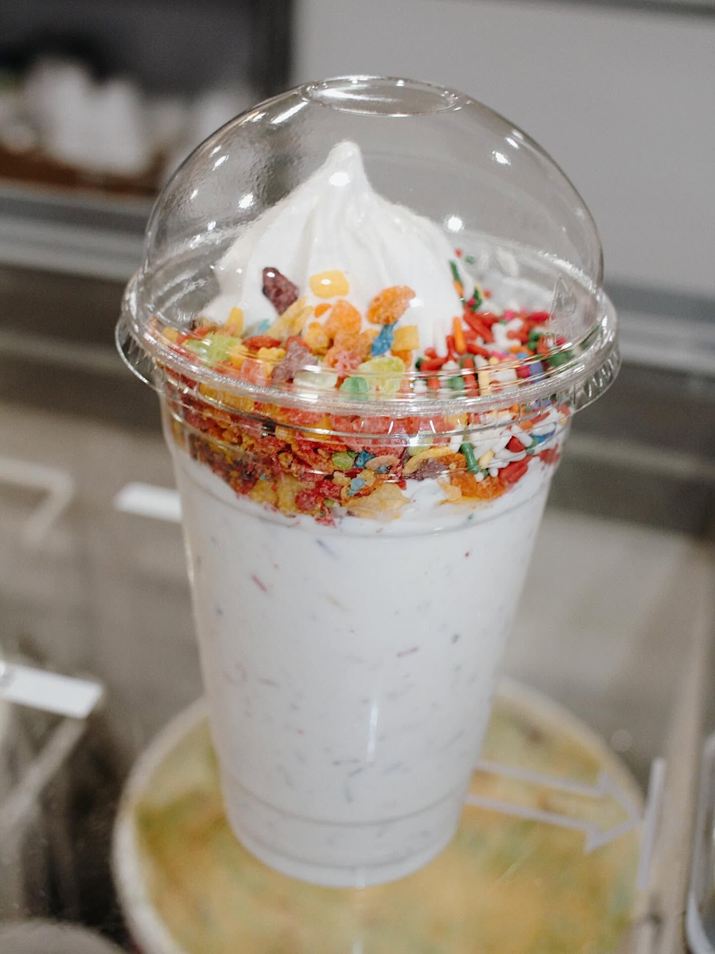 White dessert cup topped with whipped cream and colorful cereal under a clear dome lid