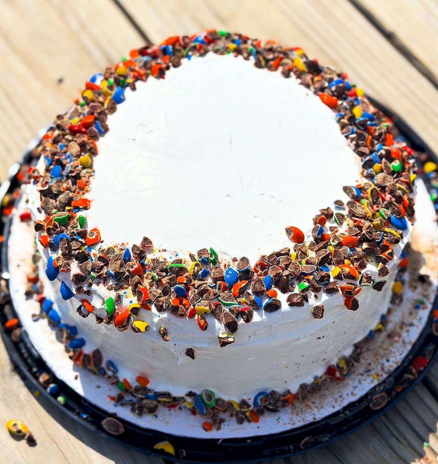 White frosted cake with colorful candy sprinkles around the top edge on a black cake board