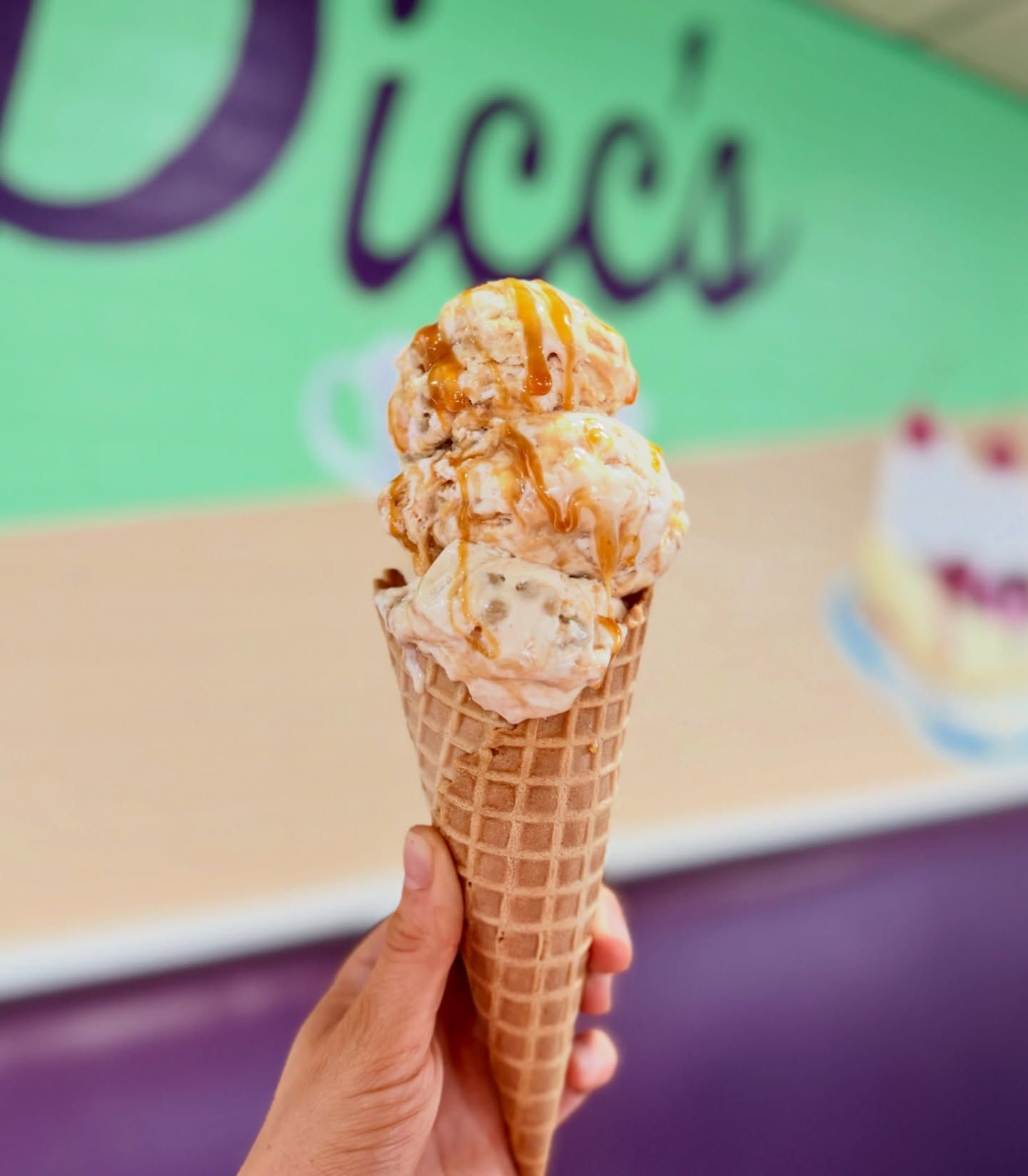 Hand holding a waffle cone with two scoops of ice cream and caramel drizzle against a green shop sign.