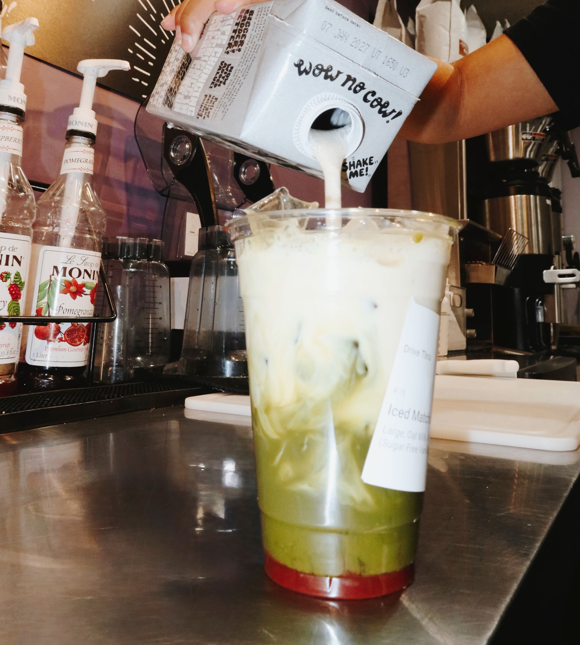 Barista pours milk into a green iced drink in a clear plastic cup at a café counter.