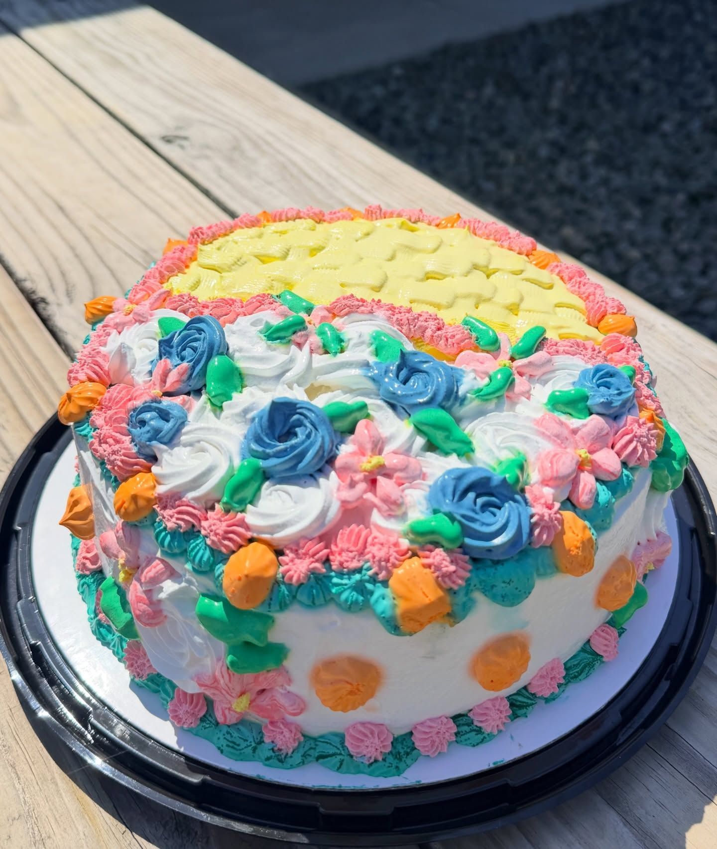 Decorated round cake with pink, yellow, green, and purple frosting flowers on a black tray outdoors