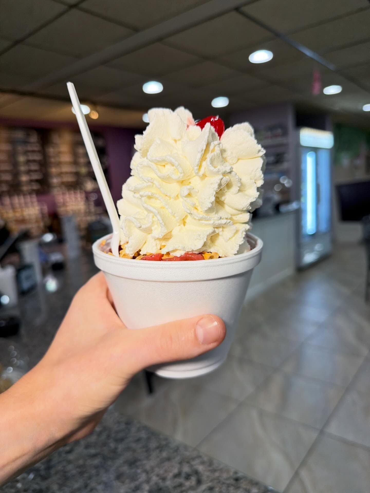 Hand holding a cup of vanilla soft-serve with strawberry sauce in a bright indoor food court