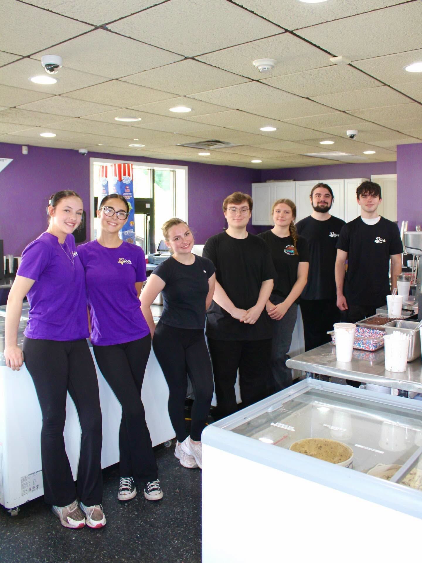 Team members in purple and black uniforms standing behind a cafeteria counter with food trays