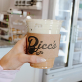 Hand holding a clear iced coffee cup with a brown sleeve labeled “Picc’s” in a café.