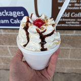 Handheld cup of a sundae topped with whipped cream, chocolate drizzle, and a cherry