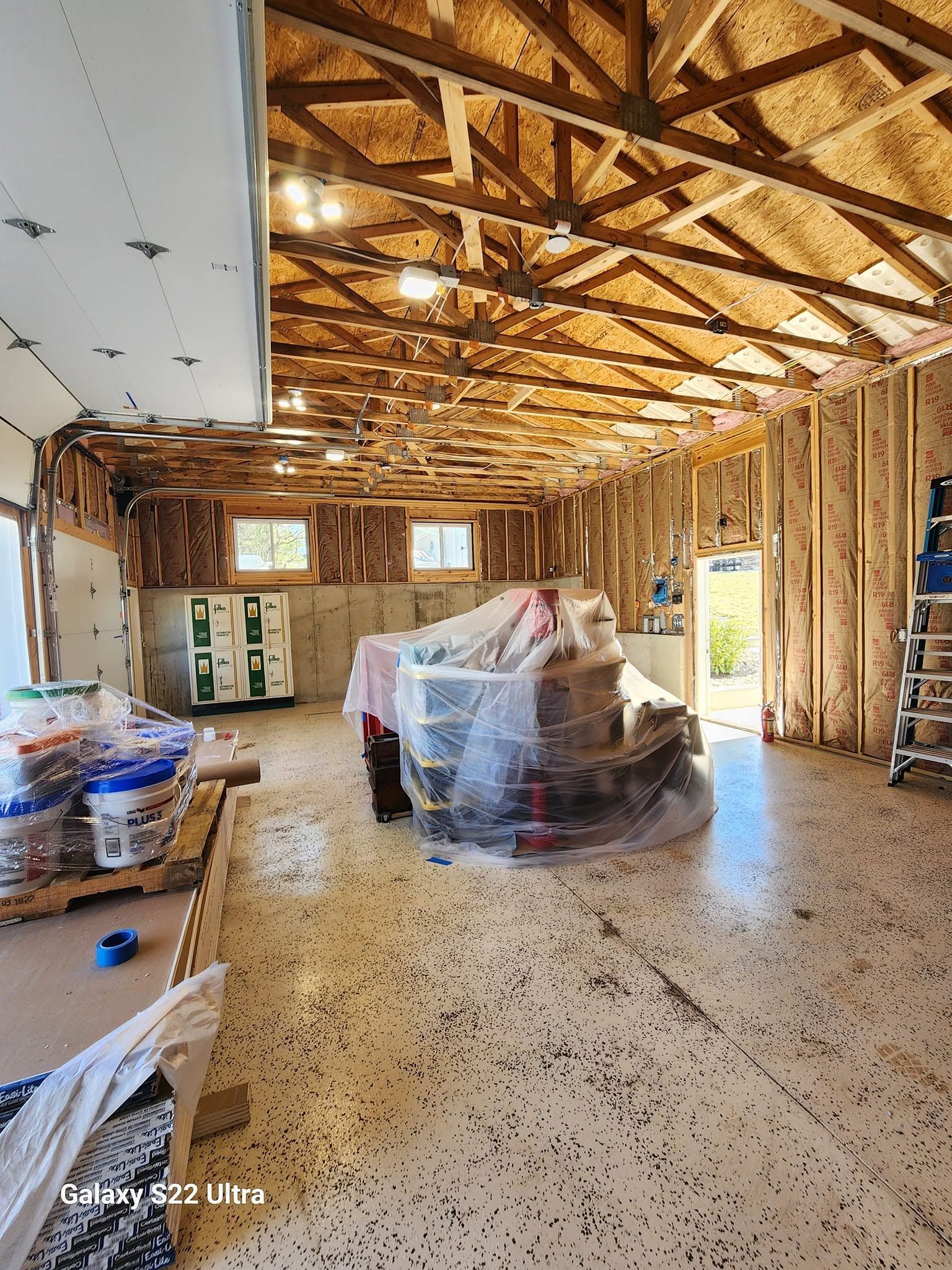 Unfinished house interior with exposed roof framing, insulation, and furniture covered in plastic over speckled floor