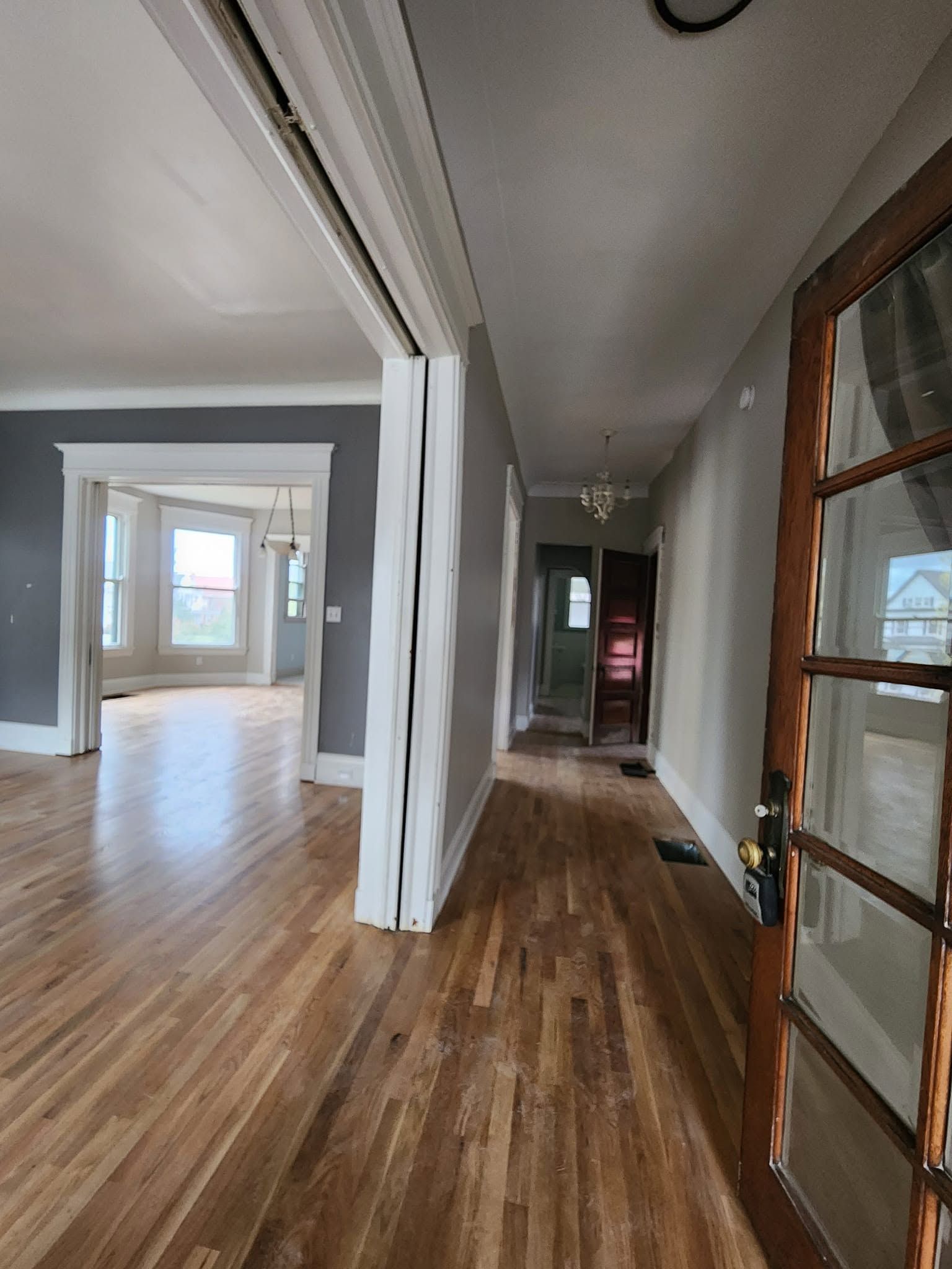 Narrow hallway with wood floors, white walls, and glass-paneled doors leading into a bright room.