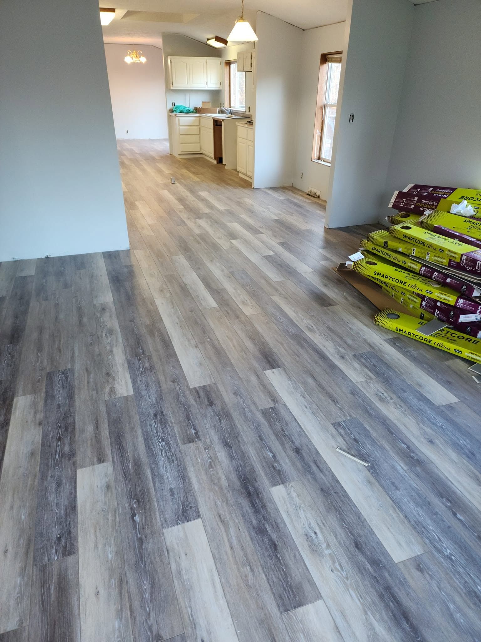 Empty living room with gray wood floors, open kitchen, and stacked flooring materials on the right.