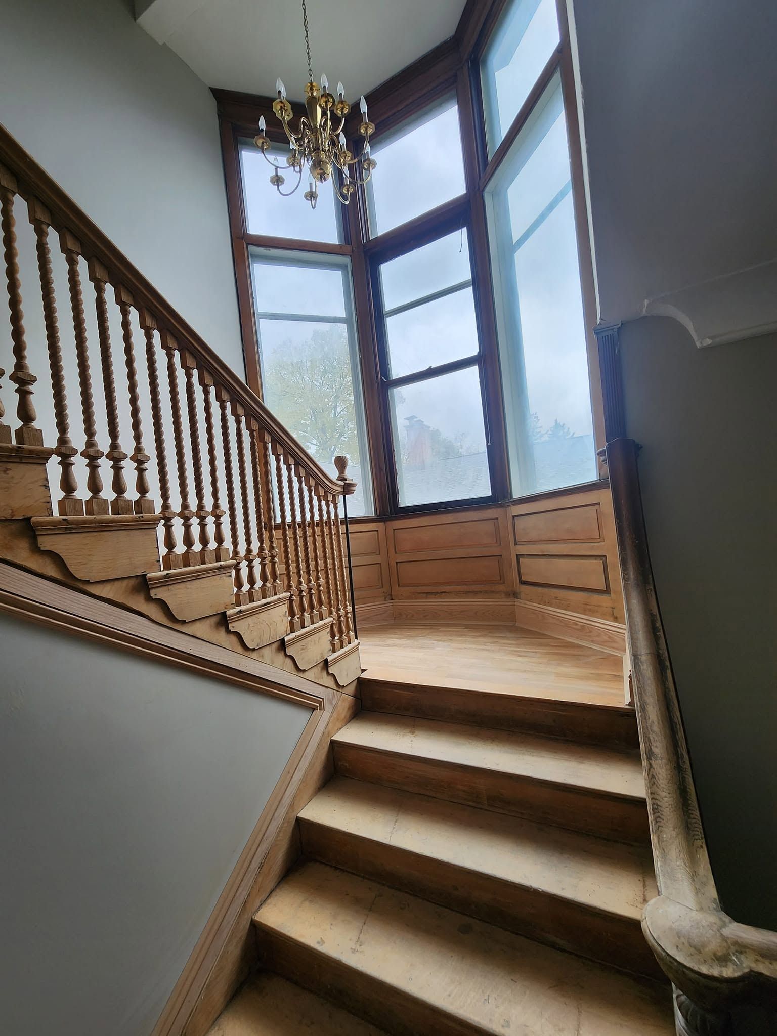 Interior staircase with wooden banister, large window, and chandelier lit by daylight