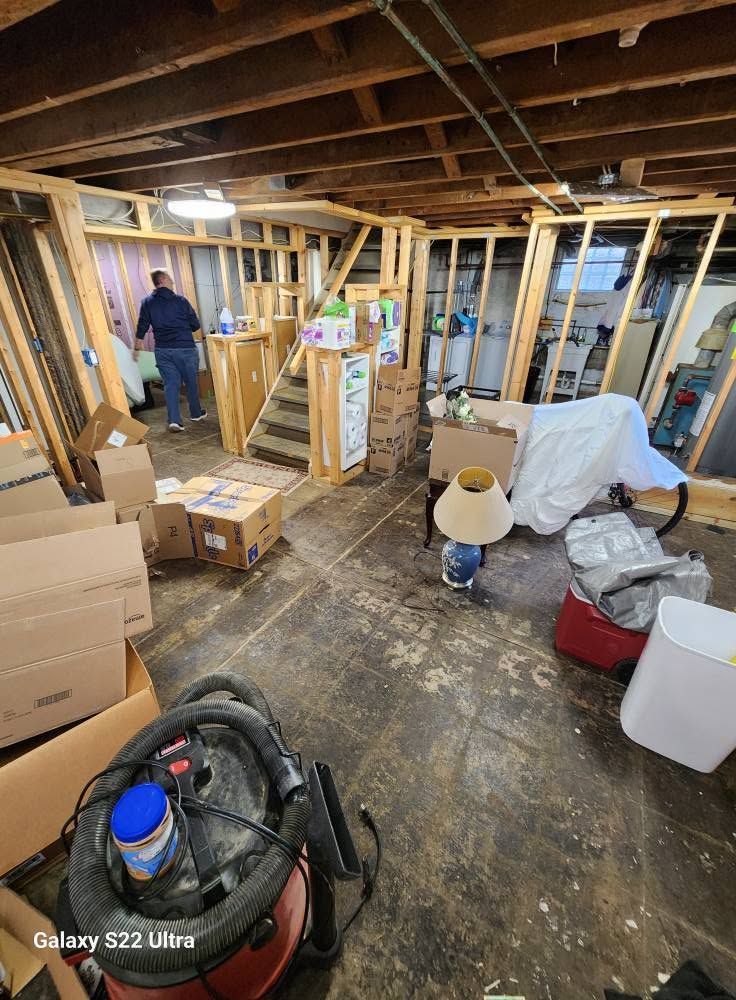 Basement renovation with exposed framing, boxes, tools, and a worker in the background.