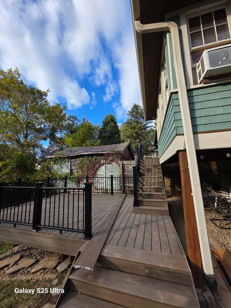 Wooden deck with black railing beside a teal house, stairs, and a sunny yard with trees and blue sky.