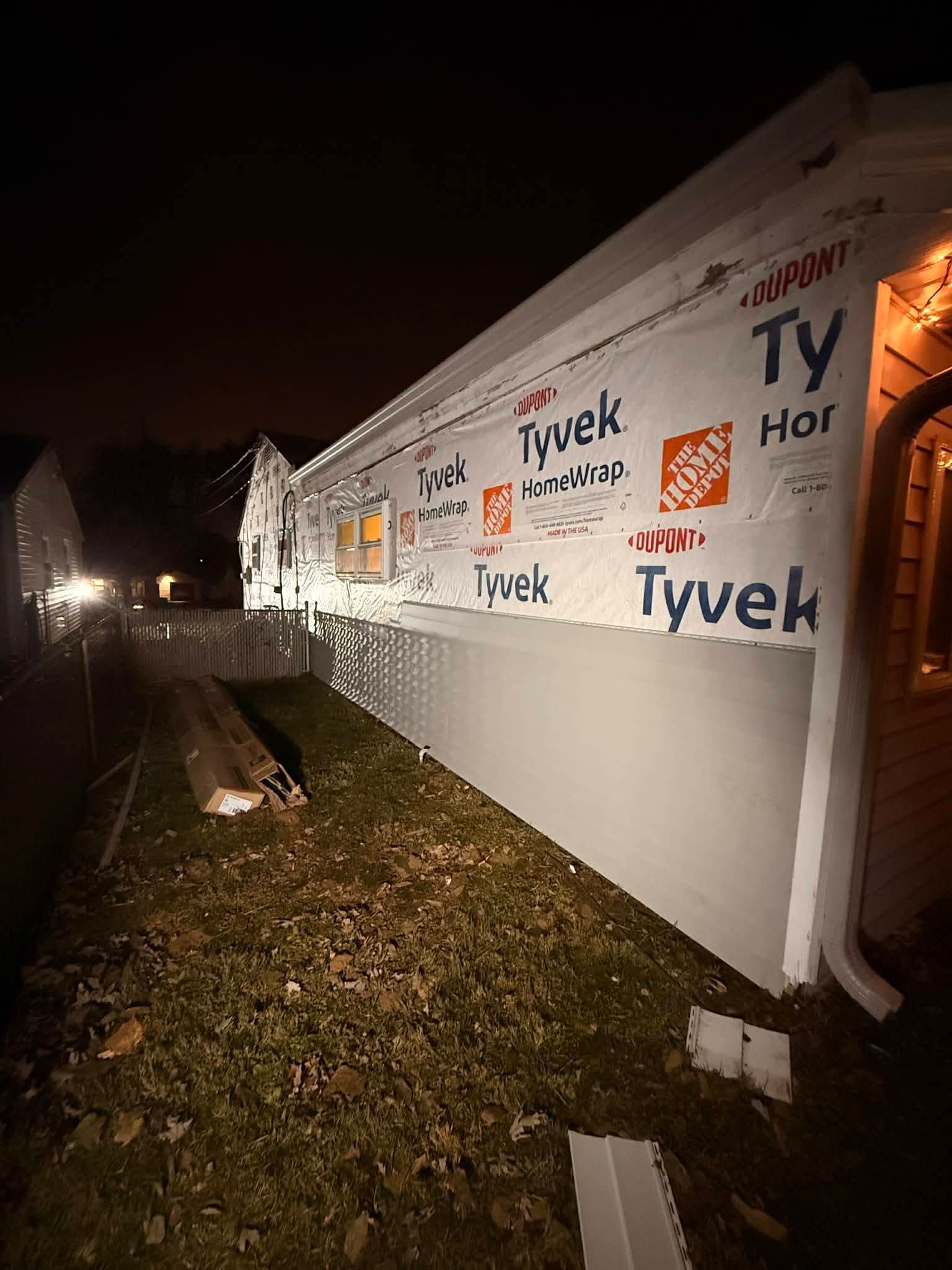 Nighttime construction site with Tyvek-wrapped building, lit by floodlights, beside a wet grassy path.