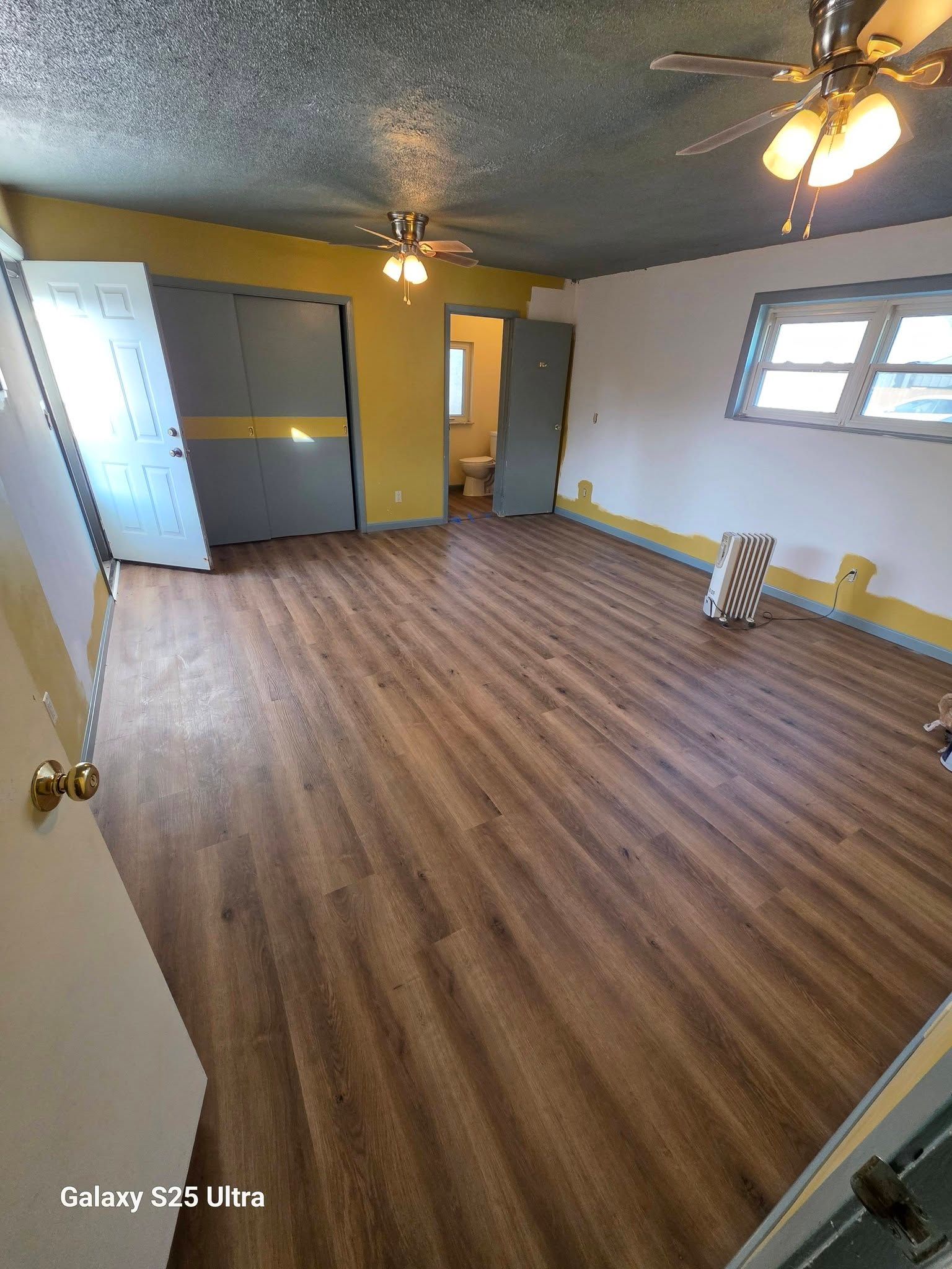 Empty room with wood-look floor, ceiling fans, white walls, and a window with a small step stool nearby.