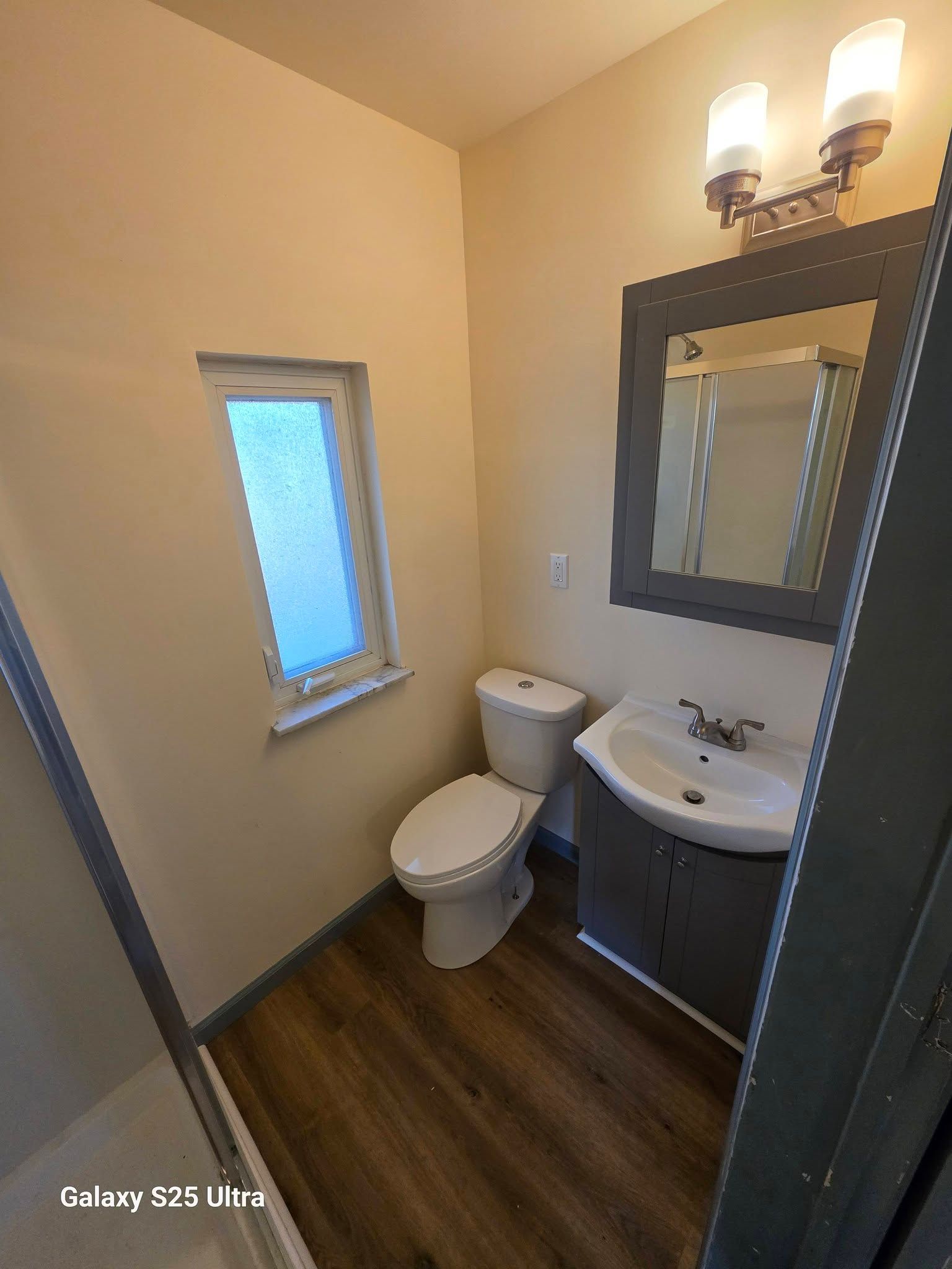 Small bathroom with toilet, sink, mirror, frosted window, and wood-look floor.