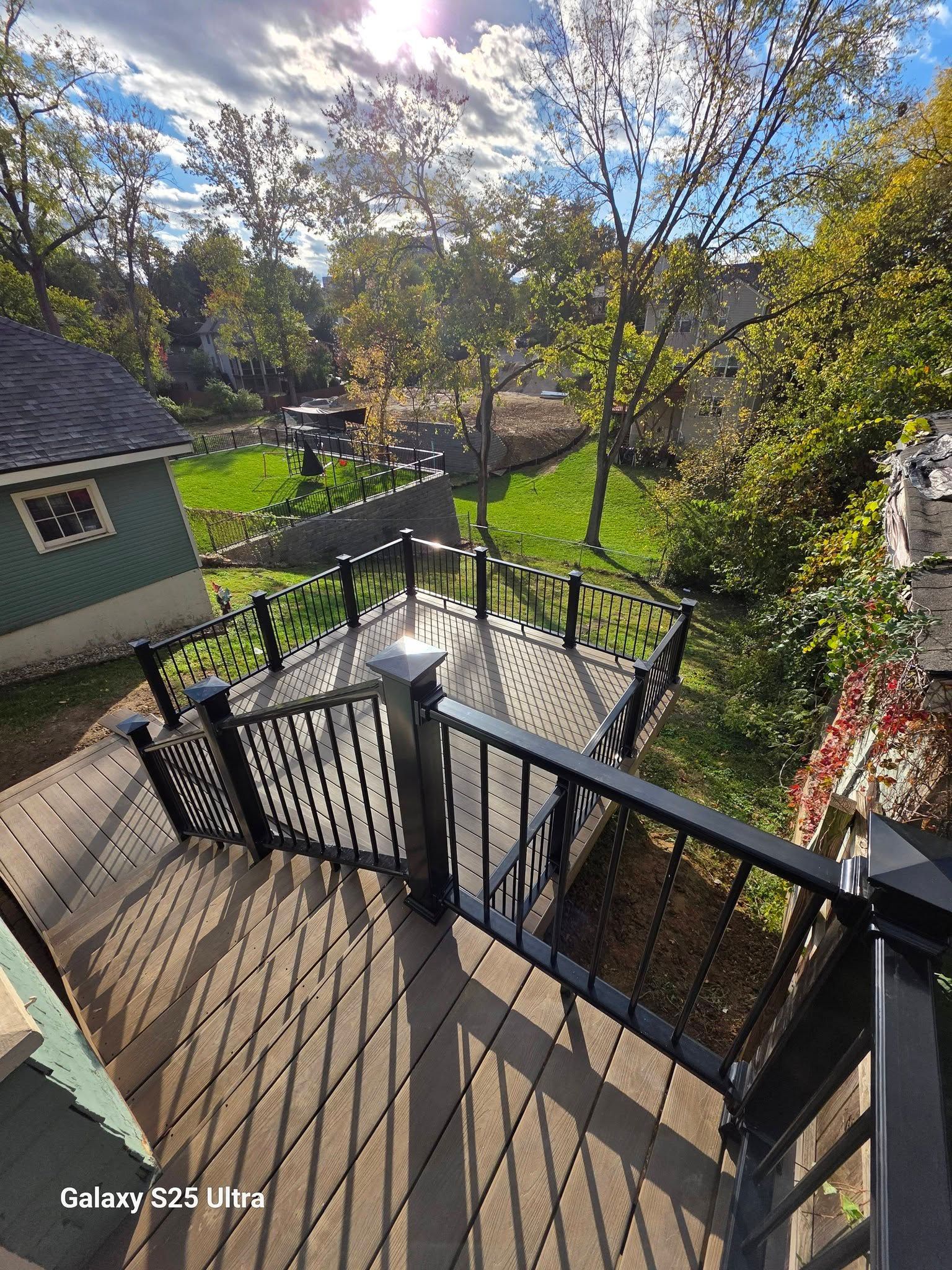 Wooden deck stairs and railings overlooking a sunny green backyard with trees and a small shed