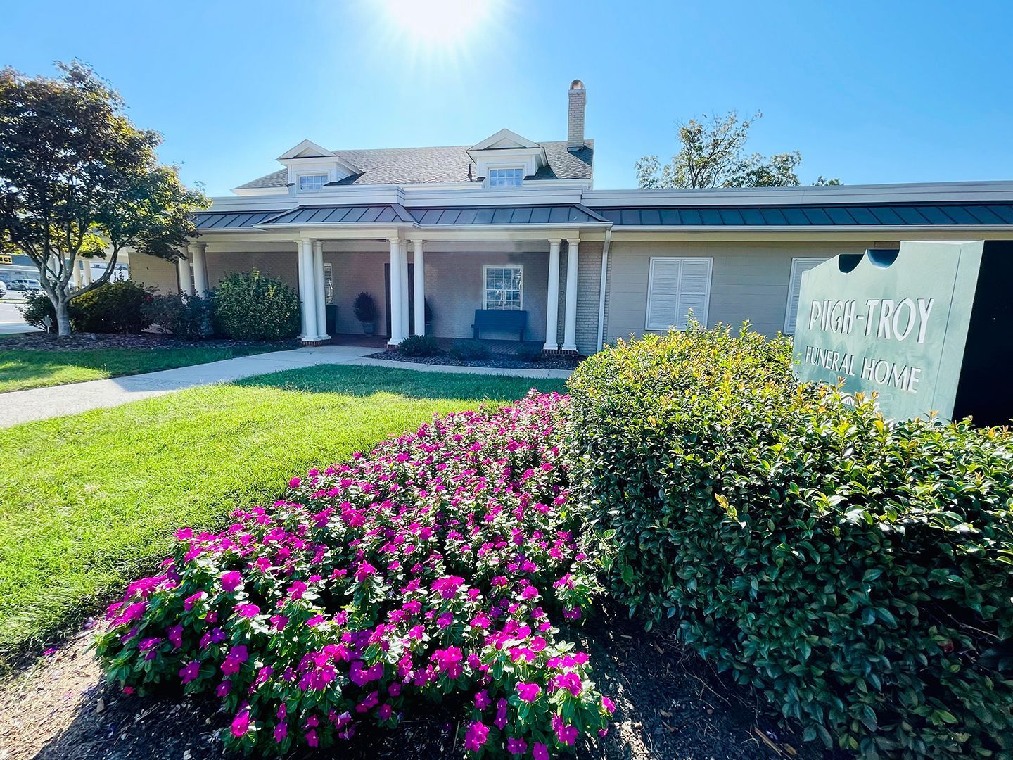 Single-story building with white columns, green lawn, pink flowers, and a sign in front under a bright sky