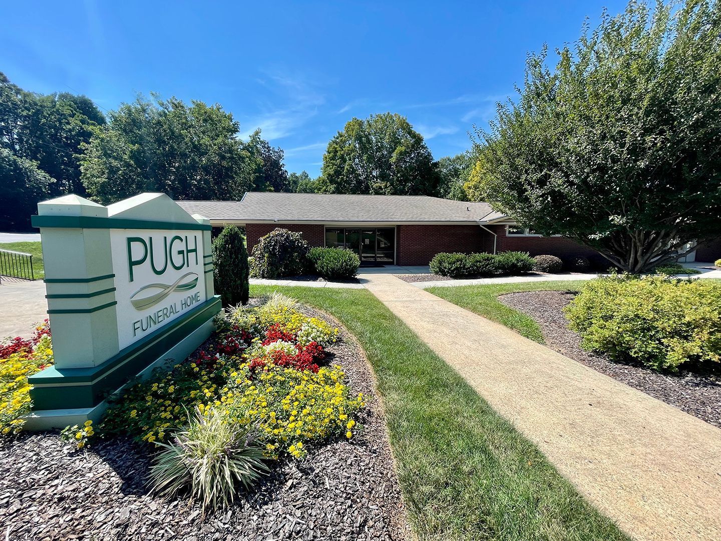 PUGH entrance sign in a landscaped garden with flowers, shrubs, and a brick building under a blue sky