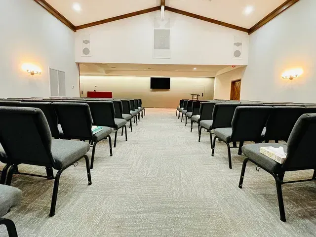 Empty meeting room with rows of black chairs facing a wall-mounted screen and podium in a bright carpeted hall