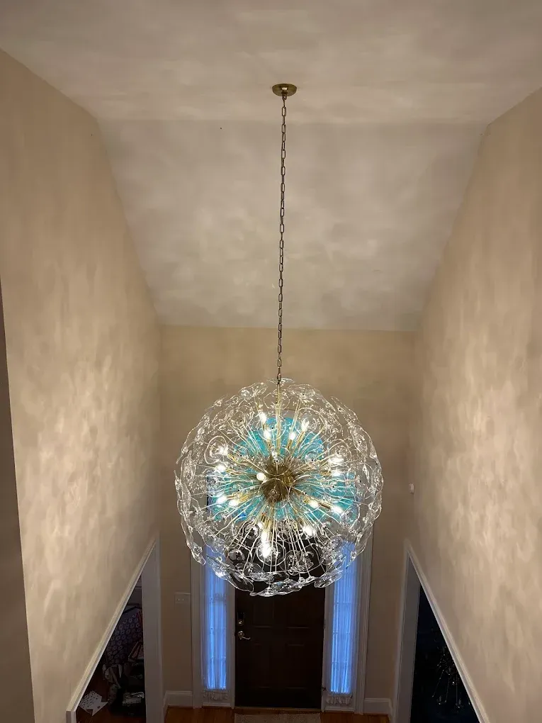 A large, ornate chandelier hangs in a tall foyer. Beige walls frame the space, with a dark door at the end.