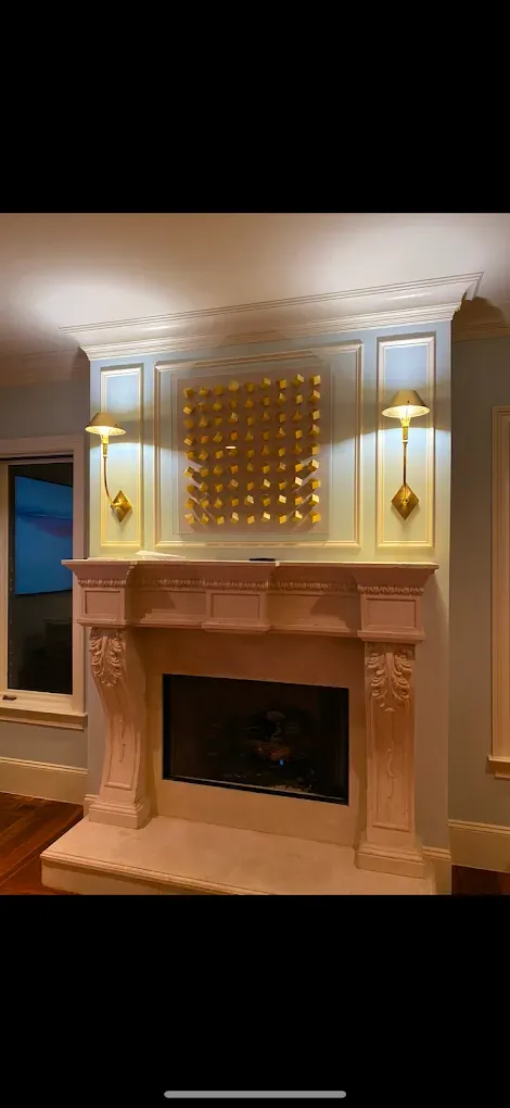 Fireplace with a gold-toned art piece, flanked by wall sconces. Ornate trim and a carved mantel.