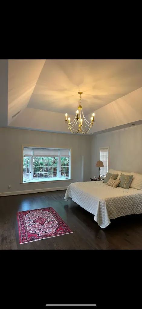 A bedroom with a dark wooden floor, large window, rug, bed, and chandelier.