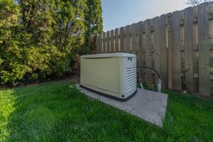 A beige home generator sits on a concrete pad in a grassy yard, next to a wooden fence.