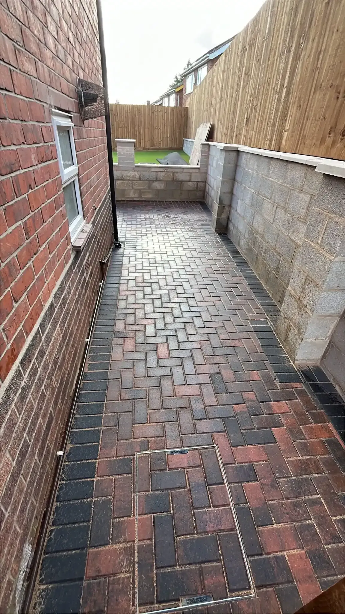 Narrow brick side alley with a wet herringbone path, stone wall, and wooden fence at the end.