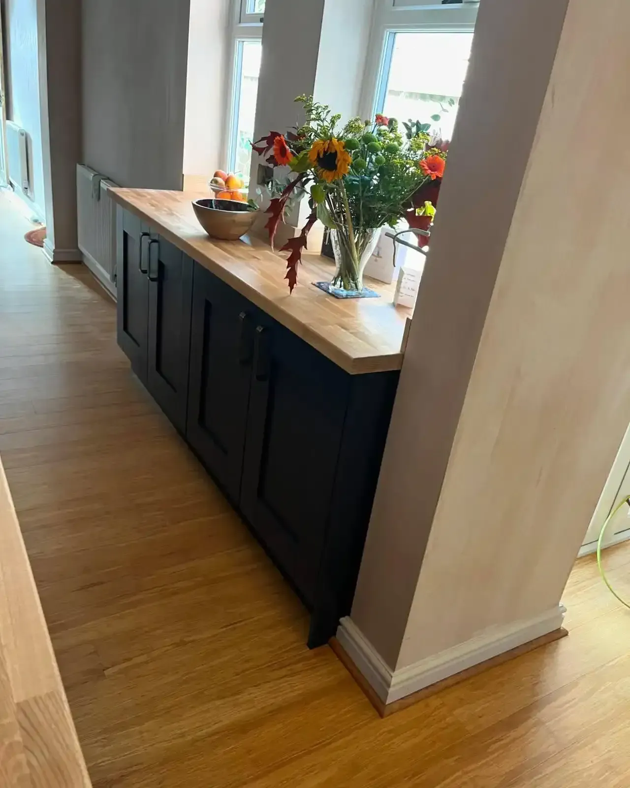Hallway console table with flowers and a bowl by a bright window, beside a hardwood floor.