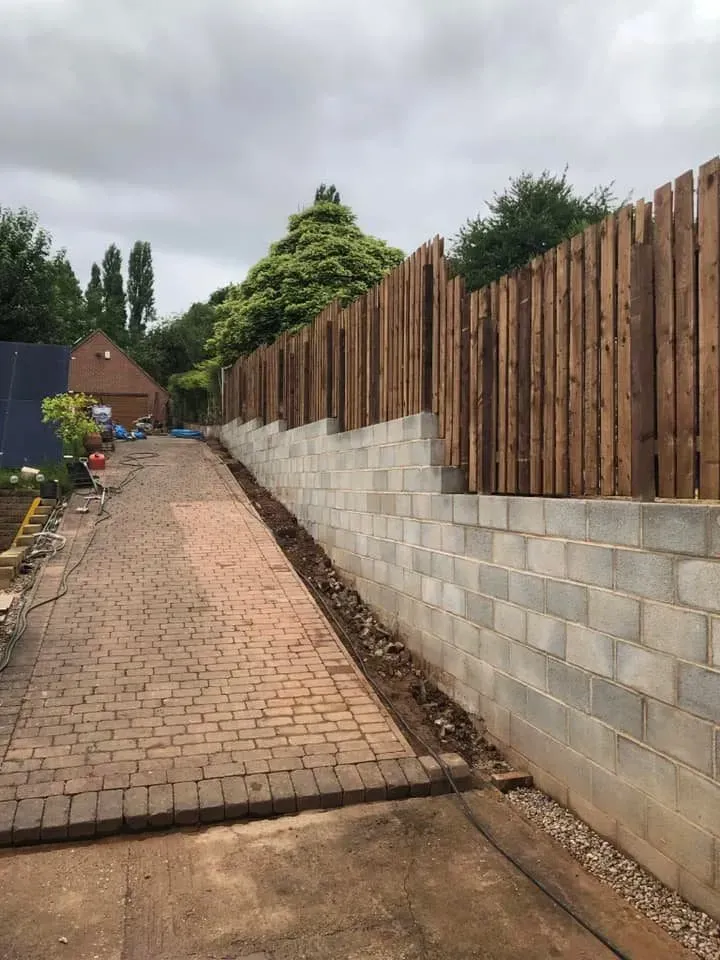 Brick driveway beside a concrete wall topped with a wooden fence, leading to houses and trees.