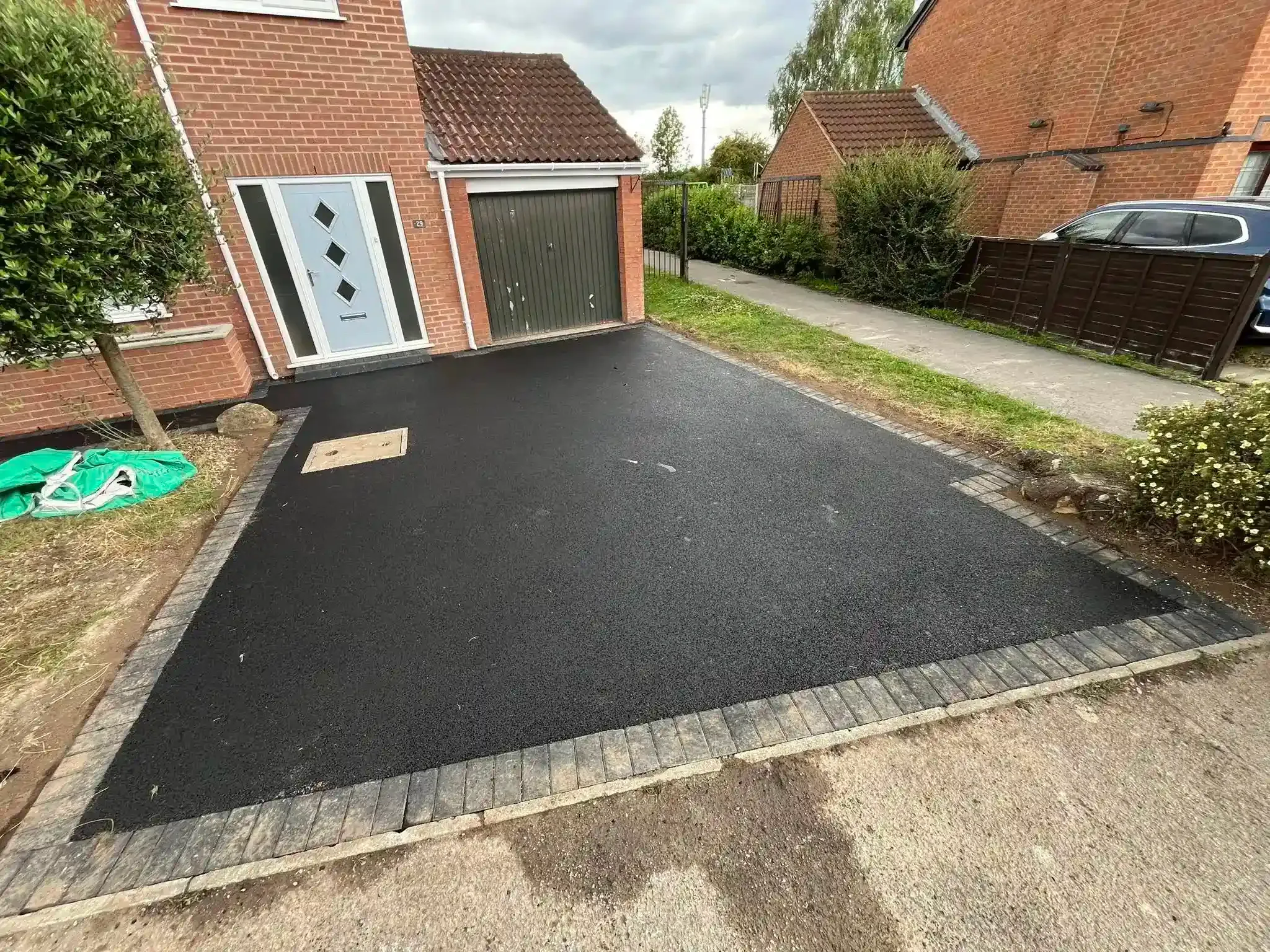 Freshly paved black driveway beside brick houses and a garage, with a narrow road and hedge on the right