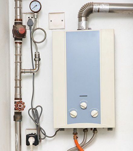 A wall-mounted gas water heater with control knobs, connected to metal piping, a pressure gauge, and power outlets.