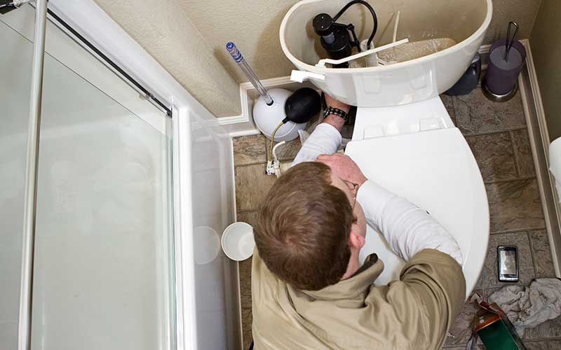 A person in a beige shirt leans over a toilet in a bathroom, reaching inside the open tank to perform repairs.