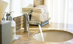 Water flows from a shiny silver faucet into a marble bathroom sink next to toiletries and a stack of folded towels.
