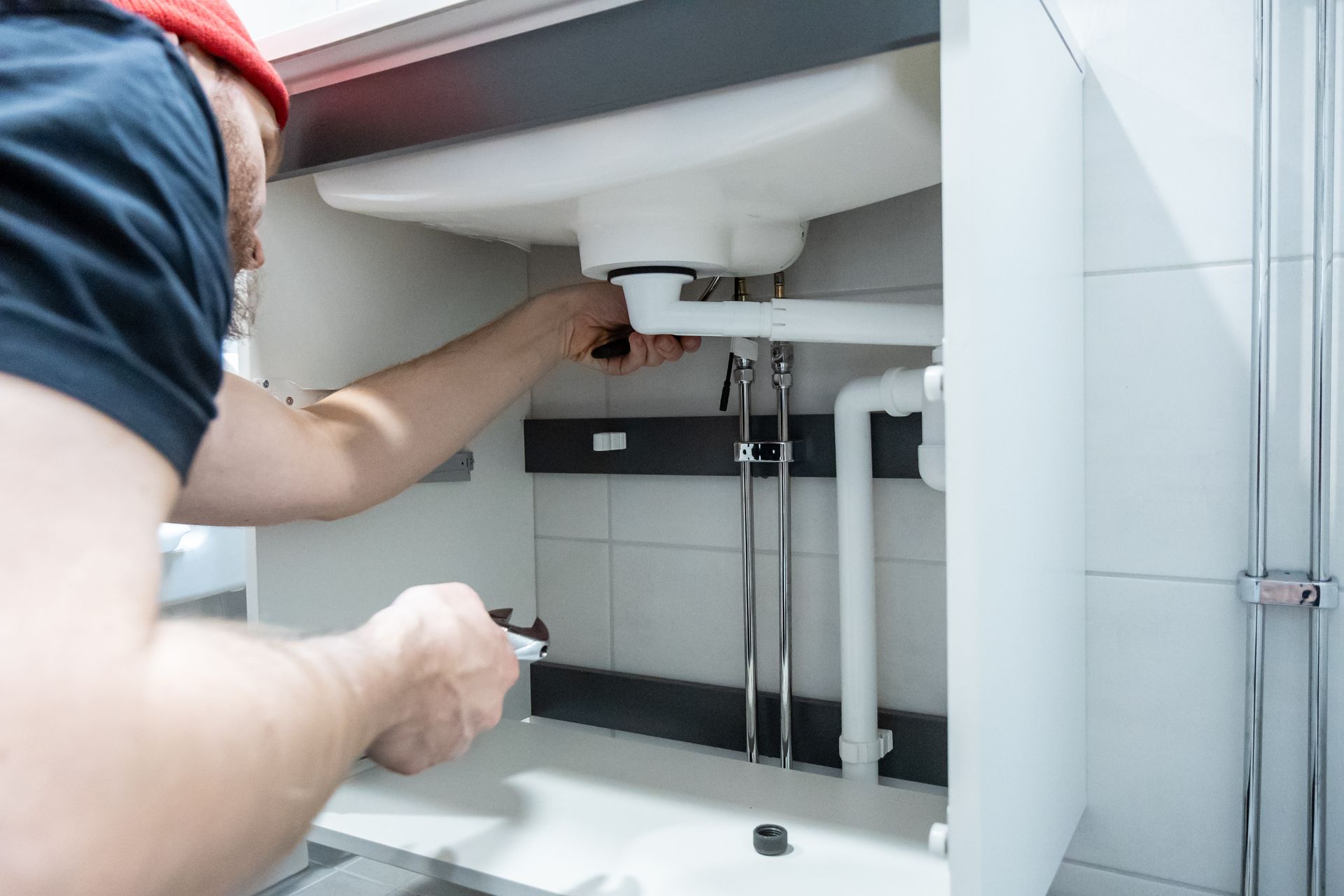 Person repairing plumbing under a white sink cabinet, hands working on exposed pipes.