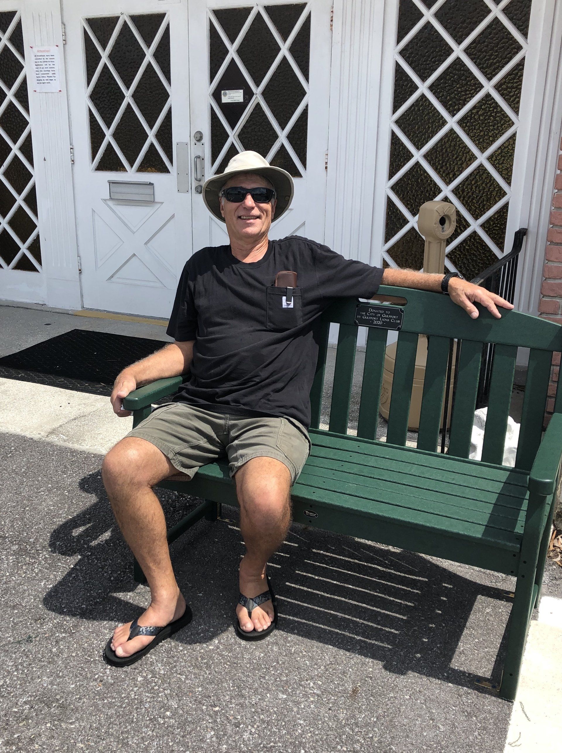 Man in hat and shorts sits on a green bench outside a building with diamond-paned windows.