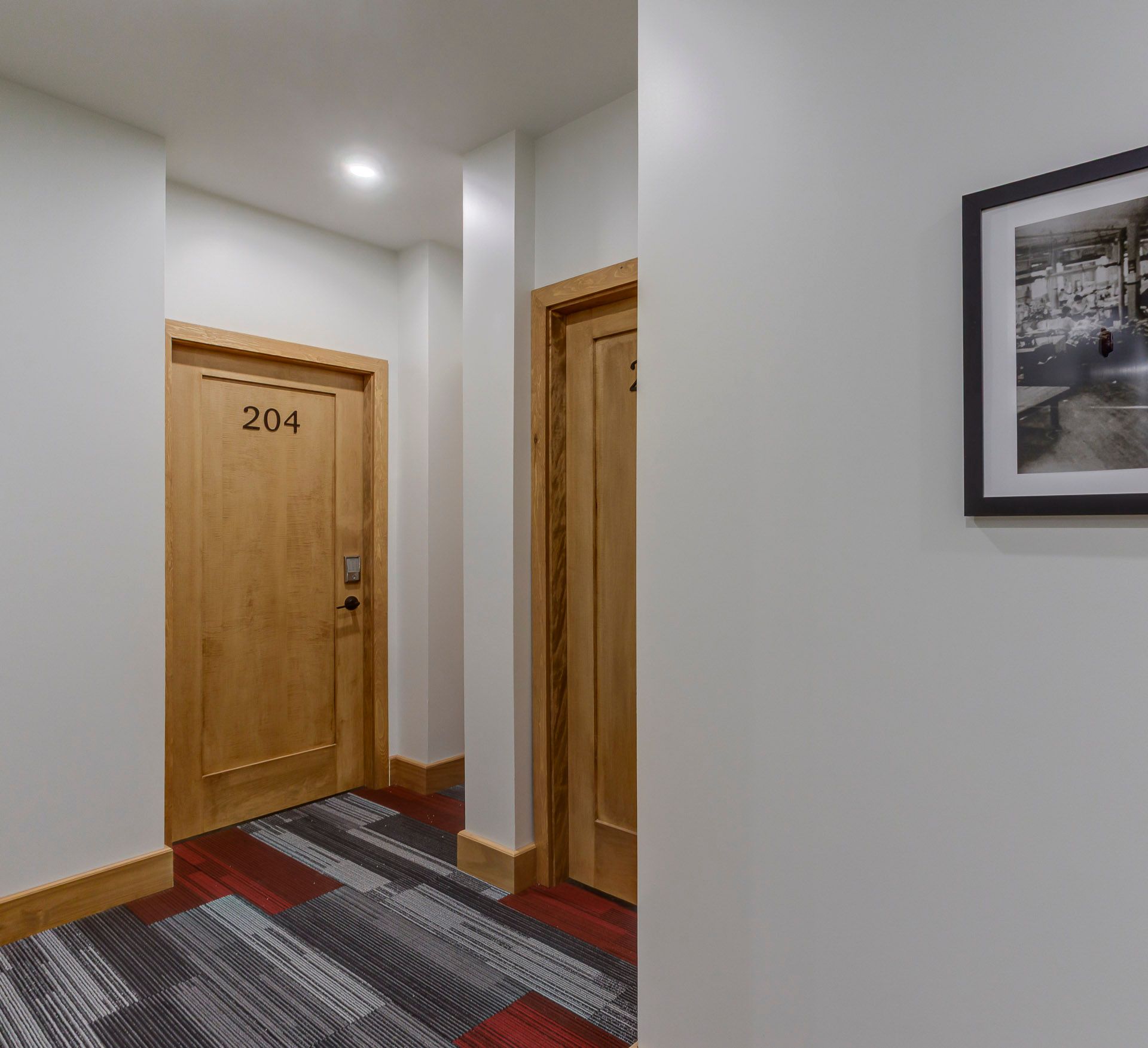 Couloir avec portes en bois, moquette à motifs et tableau encadré sur un mur blanc.