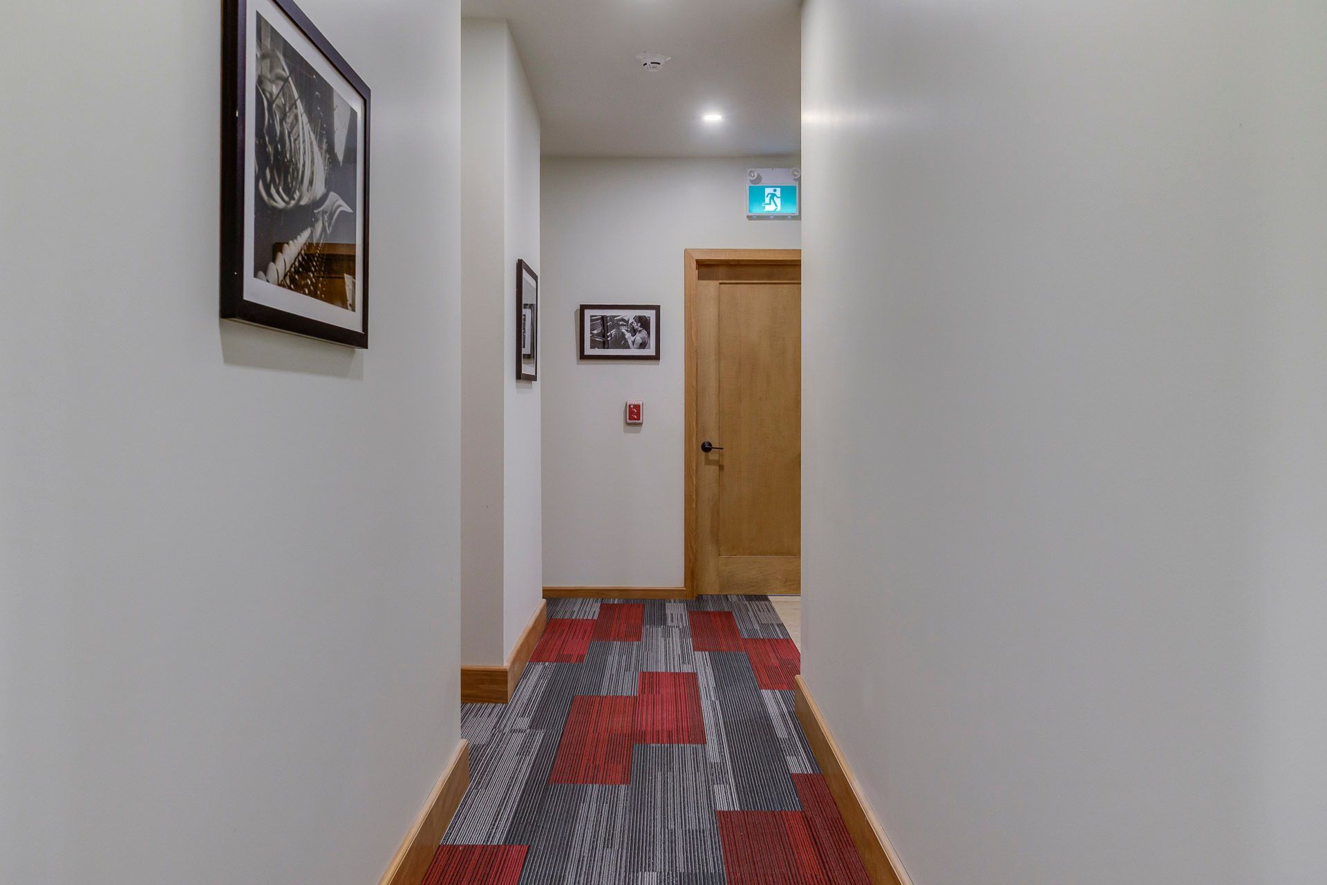 Couloir étroit aux murs blancs, moquette à motifs rouge-gris, porte en bois et tableaux encadrés.