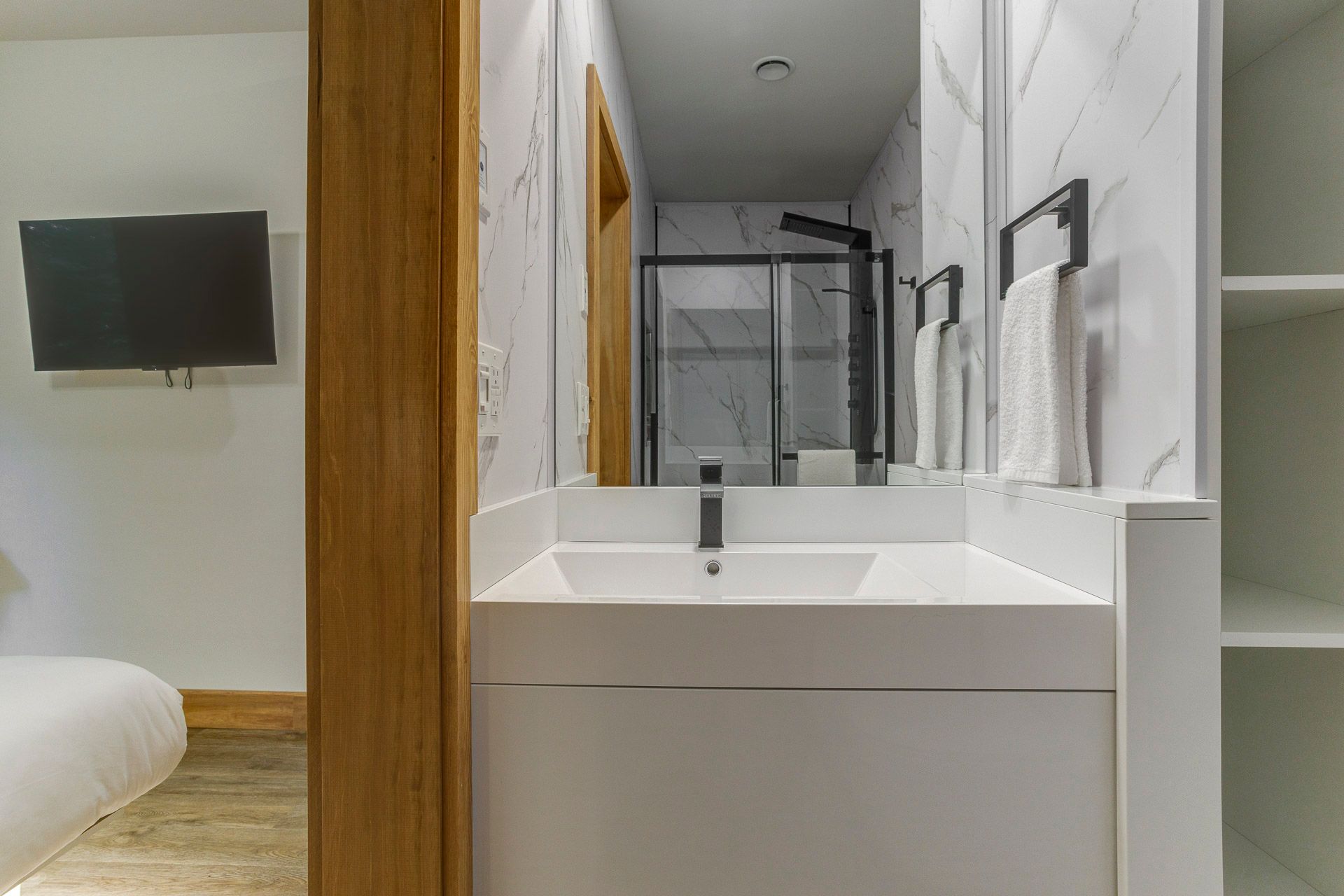 Meuble-lavabo moderne avec miroir, lavabo, porte-serviettes et aperçu d'une chambre avec télévision murale