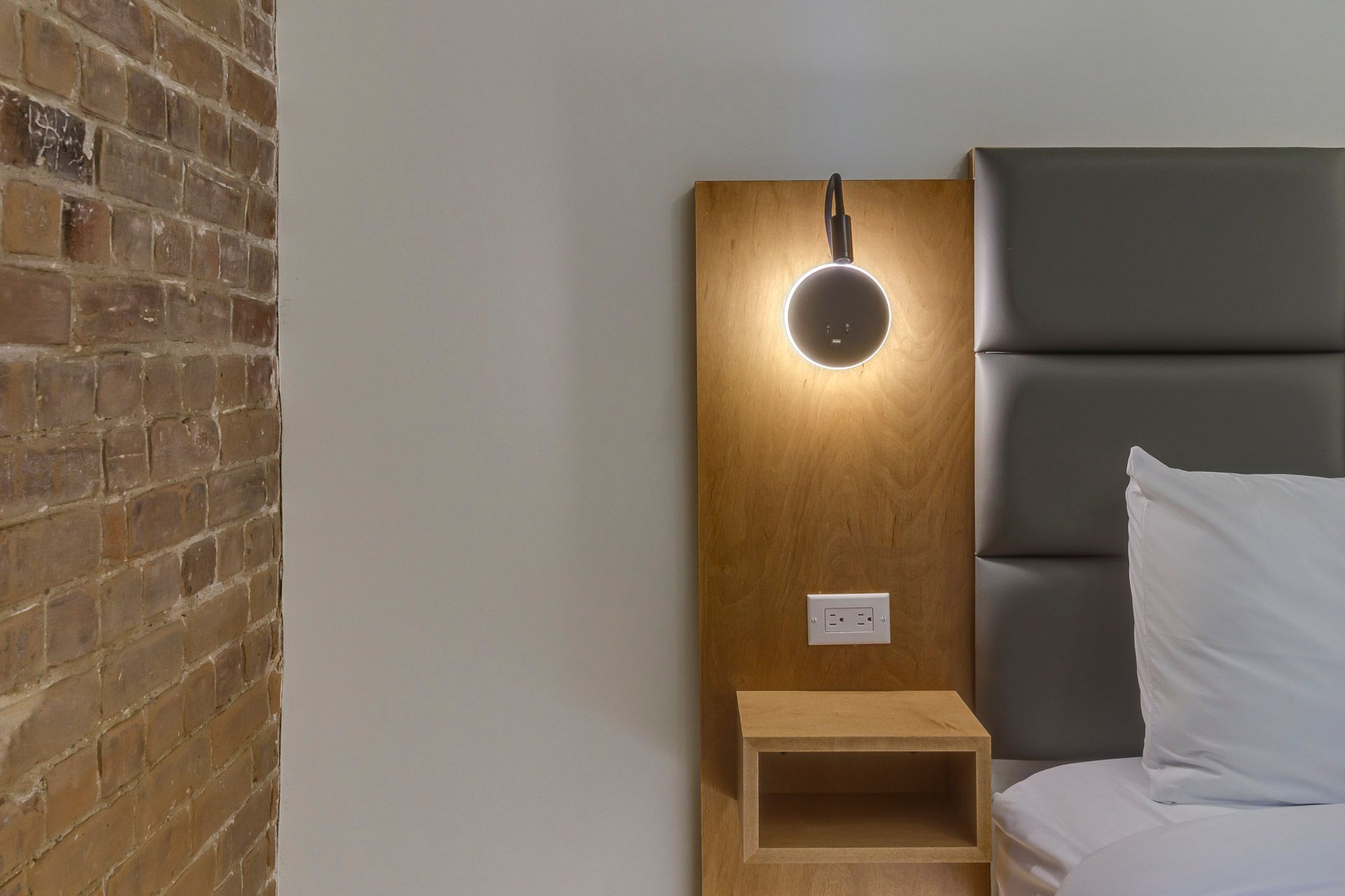 Coin lit moderne avec panneau en bois, applique murale ronde et tête de lit capitonnée grise.