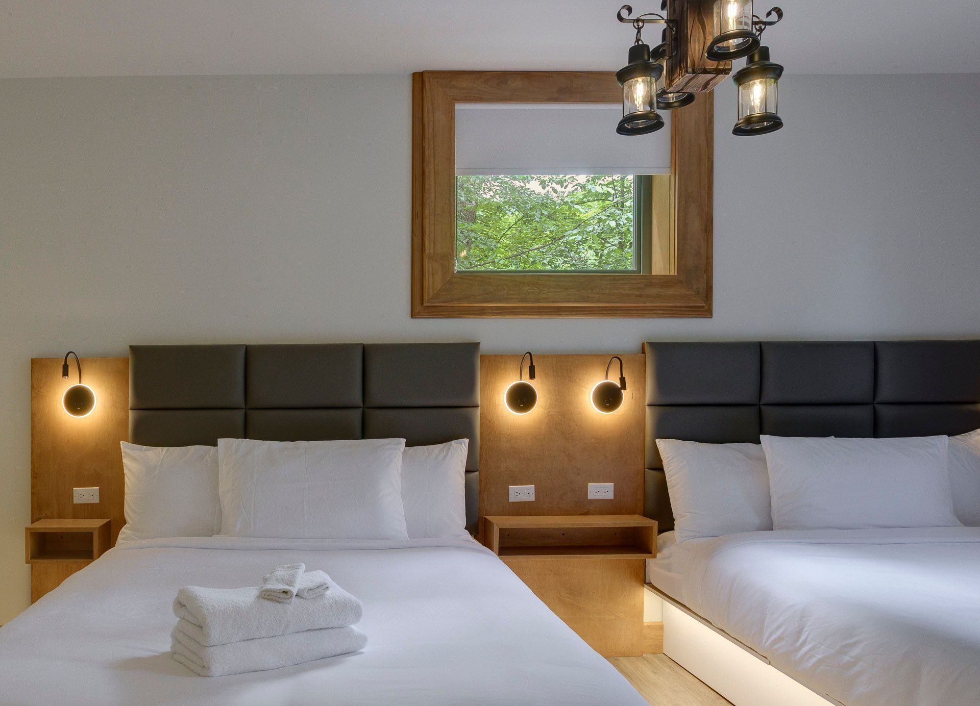 Chambre d'hôtel moderne avec deux lits queen, lampes de chevet à lumière douce et fenêtre au-dessus des têtes de lit.