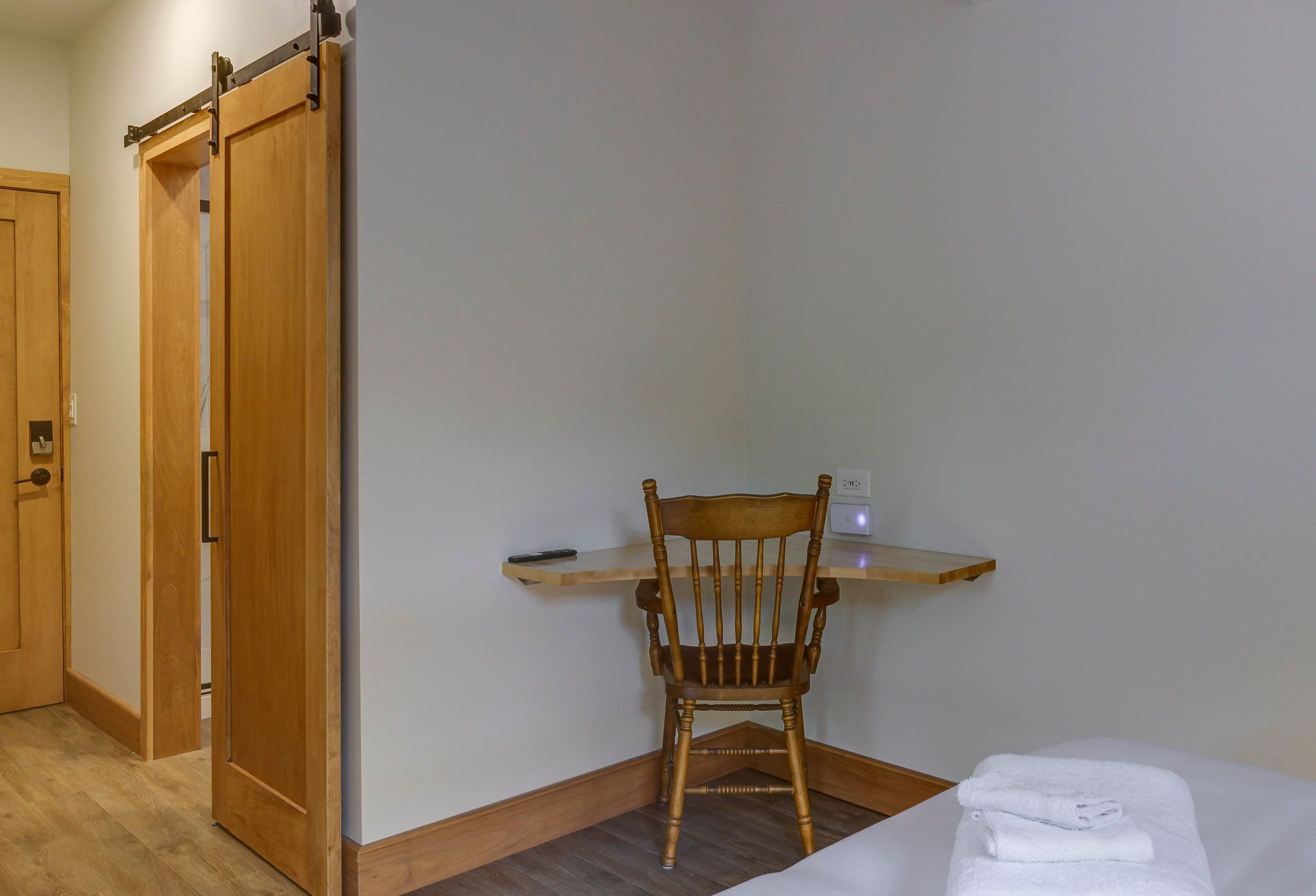 Petite chambre d'hôtel avec portes en bois, un bureau d'angle avec chaise et un coin lit avec serviettes pliées.