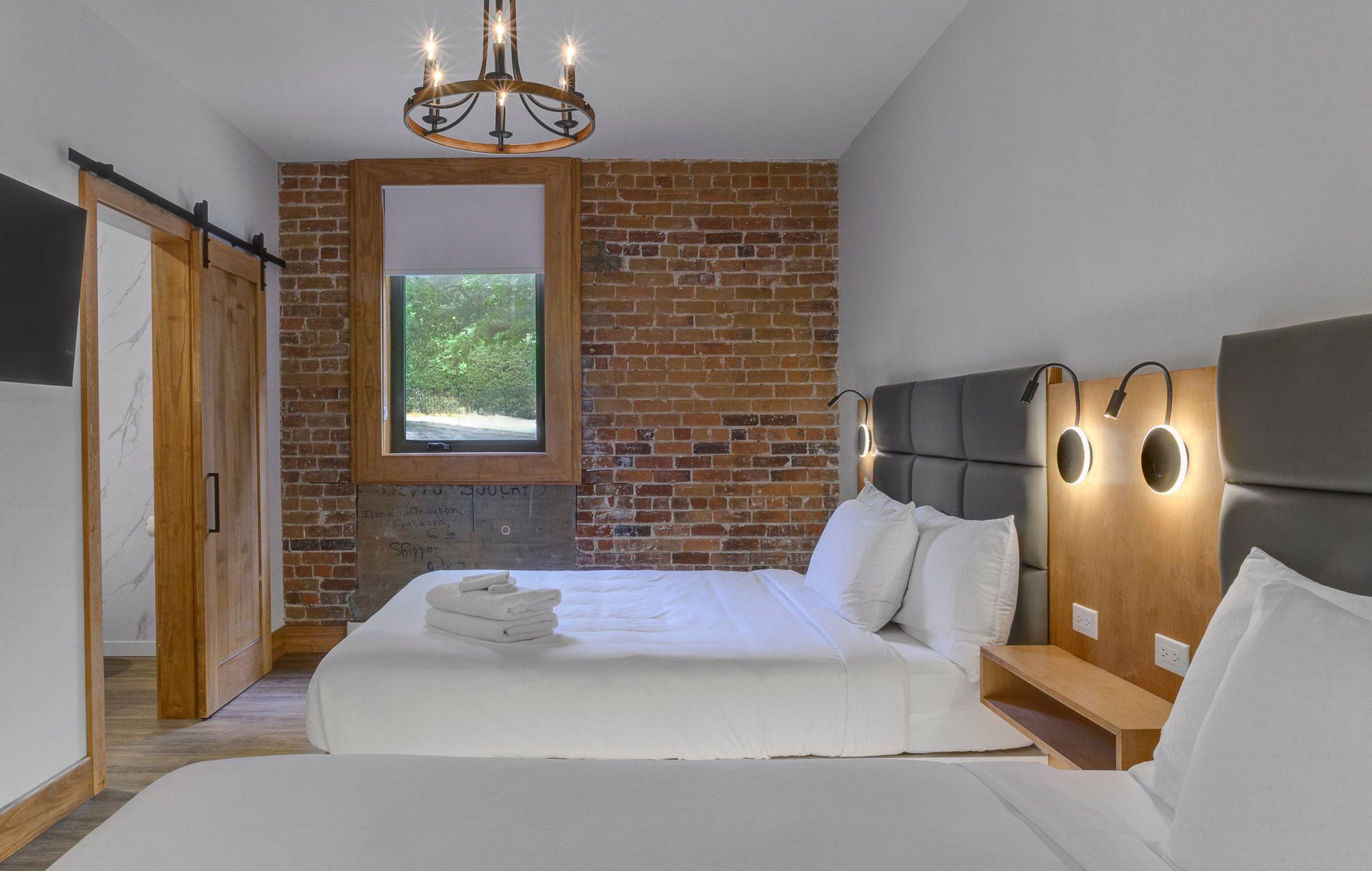 Chambre d'hôtel moderne avec mur en briques apparentes, deux lits, lustre et grande fenêtre