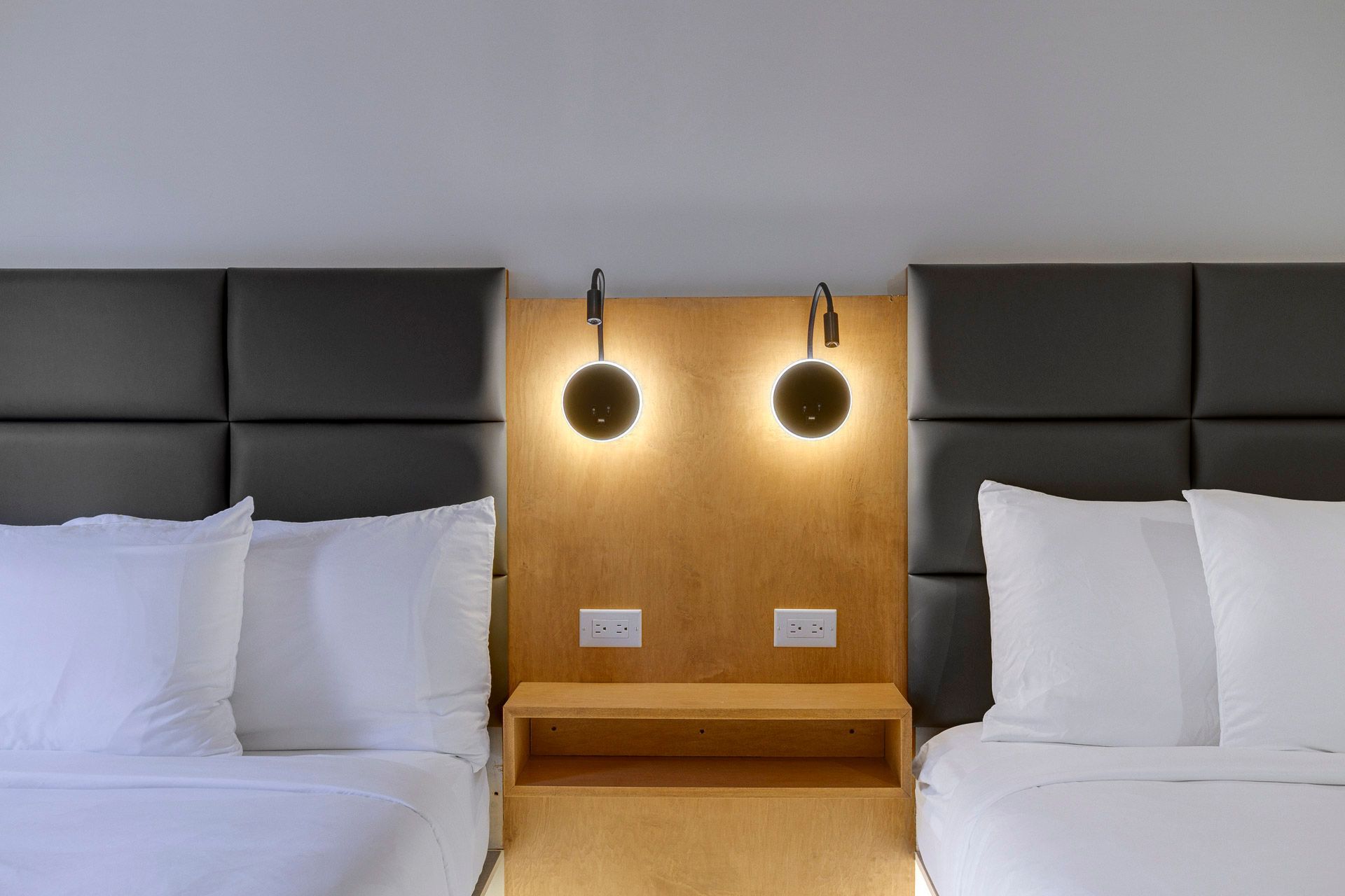 Chambre moderne avec deux lits, têtes de lit noires et panneau en bois avec deux appliques murales et prises électriques.